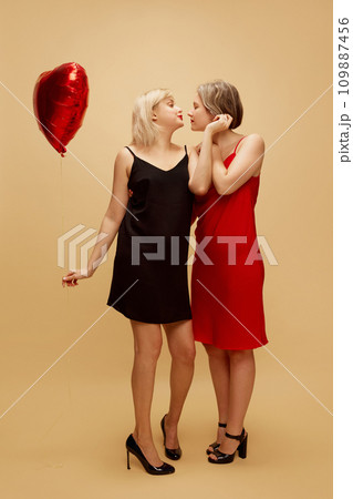 Happy young women, lesbian couple in elegant dresses celebrating romantic holiday over beige background. Valentine's Day, holiday, lgbt Happy young women, lesbian couple in elegant dresses celebrating romantic holiday over beige background. Valentine's Day, holiday, lgbt 109887456