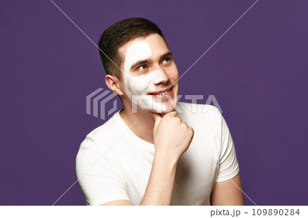 portrait of happy cheerful man looking away with of cosmetic product, white mask on face against purple background. 109890284