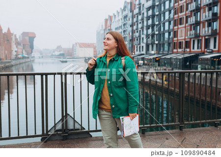 Attractive young female tourist is exploring new city. Redhead girl with backpack holding a paper map on city street in Gdansk. Traveling Europe in autumn. Famous Zuraw crane, Motlawa river Attractive young female tourist is exploring new city. Redhead girl with backpack holding a paper map on city street in Gdansk. Traveling Europe in autumn. Famous Zuraw crane, Motlawa river 109890484