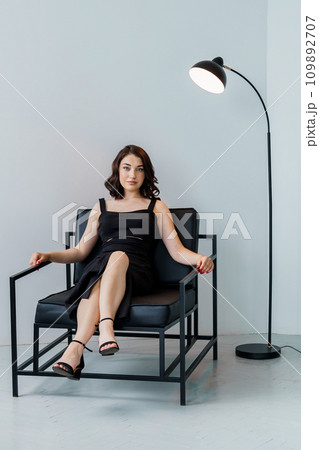 Portrait of a young beautiful girl in a black dress sitting on a chair on a gray background 109892707