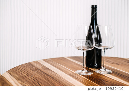 Minimalist Wine Bottle and Glasses on Wooden Table. A simple yet elegant setup of a wine bottle and two glasses on a polished wooden table against a striped background. 109894104