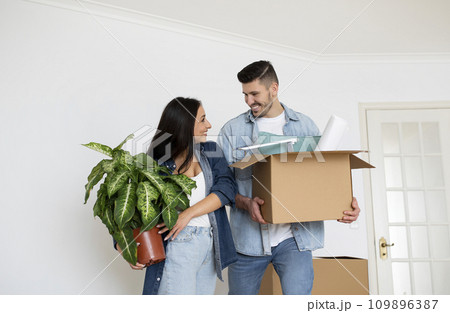 Moving Day. Cheerful Young Spouses Holding Cardboard Box And Plant In Hands 109896387