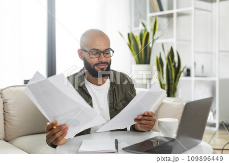 Latin male entrepreneur working from home, getting ready for presentation, sitting in front of laptop with papers Latin male entrepreneur working from home, getting ready for presentation, sitting in front of laptop with papers 109896449
