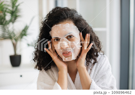 African female in white bathrobe touching face with applied paper sheet mask 109896462