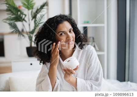 Beautiful African woman holding jar and applying cream on her face Beautiful African woman holding jar and applying cream on her face 109896613