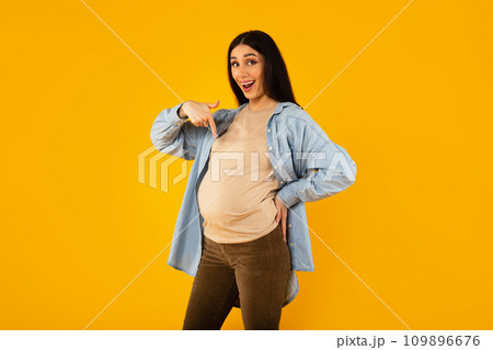 Happy pregnancy. Excited expectant woman pointing at her big belly and smiling at camera, yellow background Happy pregnancy. Excited expectant woman pointing at her big belly and smiling at camera, yellow background 109896676