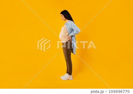 Maternity concept. Pregnant woman touching back while standing over yellow background, side view, full length shot 109896770