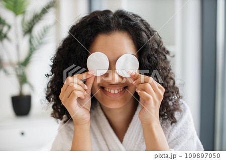 Beautiful young african female in bathrobe covering eyes with cotton pads Beautiful young african female in bathrobe covering eyes with cotton pads 109897050