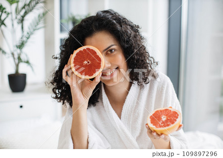 Attractive calm female in white bathrobe covering one eye with grapefruit half Attractive calm female in white bathrobe covering one eye with grapefruit half 109897296
