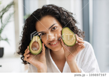 Multiracial female on robe holding two avocado halves in hands near cheek 109897397