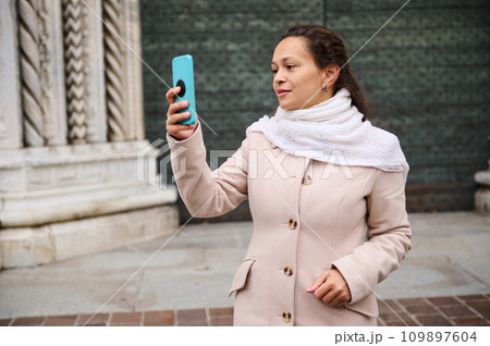 Beautiful elegant woman in beige coat, taking self portrait against medieval cathedral in Italian city in Lombardy, Como Beautiful elegant woman in beige coat, taking self portrait against medieval cathedral in Italian city in Lombardy, Como 109897604