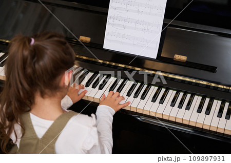 Rear view little child girl performing the rhythm of classical music while playing grand piano, putting fingers on keys Rear view little child girl performing the rhythm of classical music while playing grand piano, putting fingers on keys 109897931