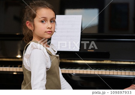 Authentic beautiful Caucasian schoolgirl looking confidently at camera, sitting at piano during individual music lesson Authentic beautiful Caucasian schoolgirl looking confidently at camera, sitting at piano during individual music lesson 109897933