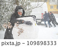 a girl is playing with snow on the street, making a snowman, on background a car is stuck in snow and men are pushing, it is snowing, blizzard and frost on a winter day 109898333