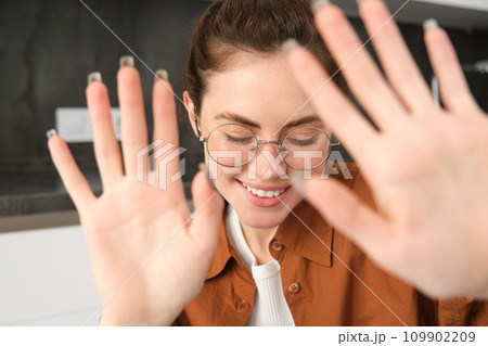 Portrait of cute and romantic young woman in glasses, covers her face, extends hands to block camera, asks to stop taking pictures Portrait of cute and romantic young woman in glasses, covers her face, extends hands to block camera, asks to stop taking pictures 109902209