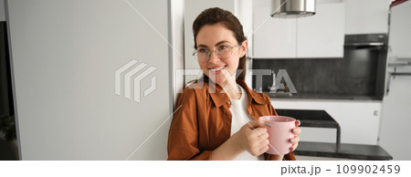 Portrait of beautiful, smiling female model, woman in glasses with cup of coffee, leans on wall at home, drinks tea 109902459