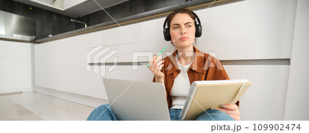 Portrait of young woman, modern student sits at home on floor and doing homework on laptop, listens music in headphones while making assignment or project for college 109902474