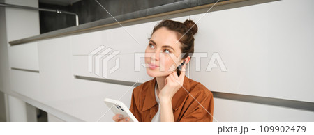 Portrait of cute young woman in wireless headphones, listening music, watching video on smartphone and using earphones, sitting on kitchen floor Portrait of cute young woman in wireless headphones, listening music, watching video on smartphone and using earphones, sitting on kitchen floor 109902479