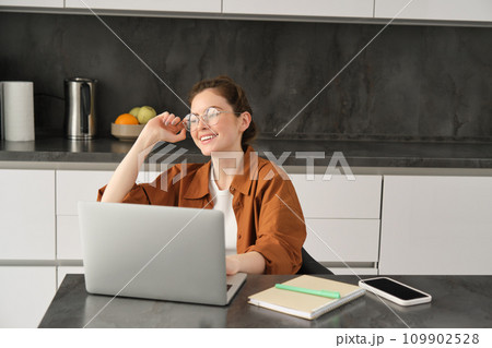 Portrait of young woman, business owner in glasses, sitting with laptop in kitchen, working from home. Student studying, doing homework on computer 109902528