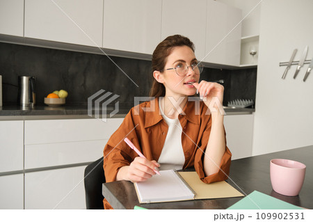 Young creative woman, writer making notes, holding pen, looking aside with thinking face, sitting in kitchen at home, doing homework 109902531