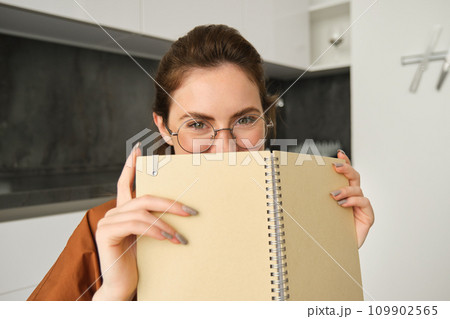 Close up portrait of cute brunette woman in glasses, sitting at home, smiling, hiding face behind yellow notebook 109902565