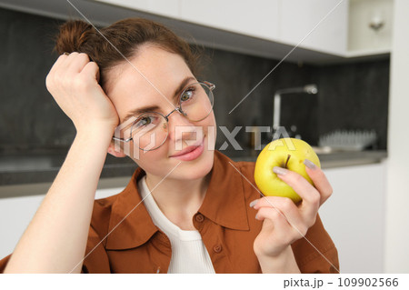 Close-up portrait of brunette woman at home, wearing glasses, eating apple in the kitchen and smiling, biting fruit 109902566