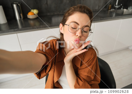 Selfie of happy young and carefree woman, taking photo on mobile phone with extended hand, posing and smiling, sitting in kitchen in glasses Selfie of happy young and carefree woman, taking photo on mobile phone with extended hand, posing and smiling, sitting in kitchen in glasses 109902628