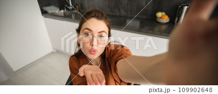 Selfie of happy young and carefree woman, taking photo on mobile phone with extended hand, posing and smiling, sitting in kitchen in glasses 109902648