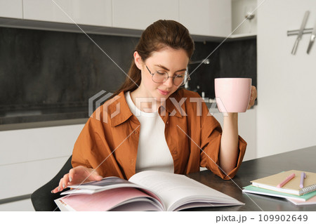 Distance learning. Young woman at home, student studying at home and drinking tea, reading her work book, revising for exam Distance learning. Young woman at home, student studying at home and drinking tea, reading her work book, revising for exam 109902649