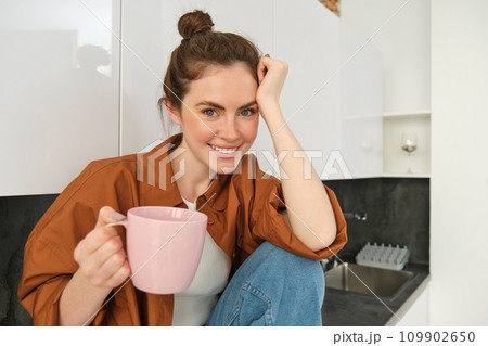 Portrait of young woman relaxing in kitchen with cup of coffee, smiling at camera, sitting on kitchen counter 109902650
