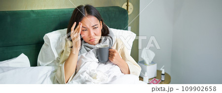 Flu, sickness and people. Young woman feeling sick, having fever and catching col, lying in bed with hot tea, taking drugs from influenza Flu, sickness and people. Young woman feeling sick, having fever and catching col, lying in bed with hot tea, taking drugs from influenza 109902658