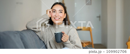 Smiling asian woman sitting at home with cup of coffee, relaxing and feeling warmth, looking outside window, resting on sofa in living room 109902706