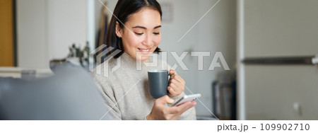 Portrait of smiling asian girl checking her news feed on smartphone and drinking coffee, sitting on sofa at home, browsing on mobile phone, reading 109902710