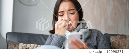 Ill korean woman sitting at home, sneezing, caught cold, reading news on mobile phone, freezing at her home because of heating problems Ill korean woman sitting at home, sneezing, caught cold, reading news on mobile phone, freezing at her home because of heating problems 109902714