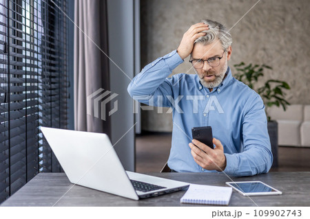Sad disappointed boss businessman at workplace received online message email on phone, senior mature gray haired man using app on smartphone, inside office with laptop. Sad disappointed boss businessman at workplace received online message email on phone, senior mature gray haired man using app on smartphone, inside office with laptop. 109902743