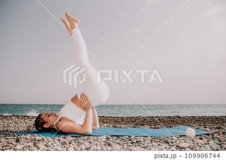 Woman sea yoga. Well looking middle aged woman with braids dreadlocks in white leggings and tops doing stretching pilates on yoga mat near sea. Female fitness yoga routine concept. Healthy lifestyle. 109906744