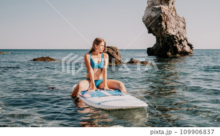 Woman sea sup. Close up portrait of happy young caucasian woman with long hair looking at camera and smiling. Cute woman portrait in a blue bikini posing on sup board in the sea 109906837