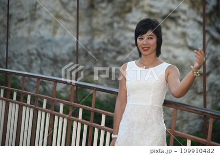 Portrait photography of a mature, beautiful, happy Asian woman. Model in white casual clothes. Standing and smiling at the camera, standing on a bridge with mountains behind. 109910482