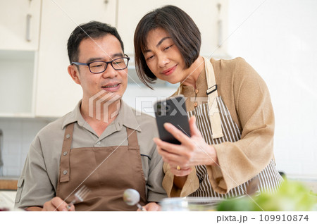 Happy Asian wife is showing something on her phone to her husband while he is eating in the kitchen. 109910874