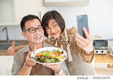 A lovely Asian couple taking pictures or selfies with their food in the kitchen together. A lovely Asian couple taking pictures or selfies with their food in the kitchen together. 109910879