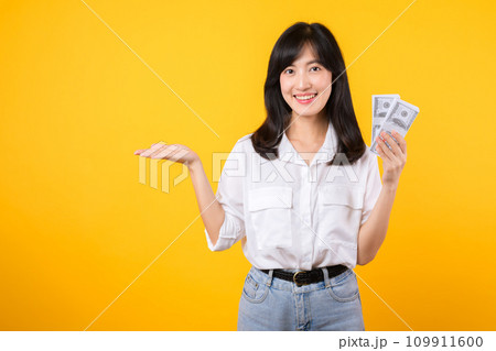Young happy woman of Asian ethnicity wear white shirt and denim jean holding cash money in dollar and pointing hand to free copy space against yellow background. Investment and financial concept. Young happy woman of Asian ethnicity wear white shirt and denim jean holding cash money in dollar and pointing hand to free copy space against yellow background. Investment and financial concept. 109911600
