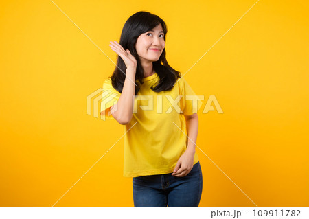 Capture attention portrait of young Asian woman. Wearing a yellow t-shirt and denim jeans, leans in to overhear and listen, evoking curiosity and intrigue. attention-grabbing promotions and discounts. 109911782