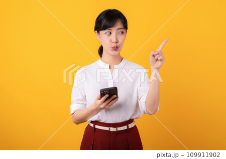 Portrait beautiful young asian woman enterpriser happy smile wearing white shirt and red plants pointing finger gesture to free copy space and using smartphone isolation on yellow background. 109911902