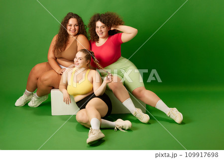 Three young women with oversized bodies sitting in comfortable sportswear against green studio background. Gym Three young women with oversized bodies sitting in comfortable sportswear against green studio background. Gym 109917698