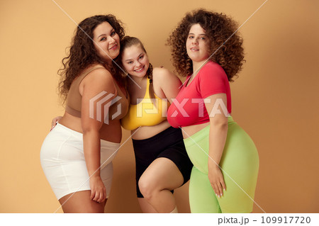 Elegant, positive, smiling young women, friends standing in sportswear over beige studio background. Oversize models Elegant, positive, smiling young women, friends standing in sportswear over beige studio background. Oversize models 109917720