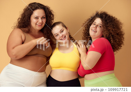 Elegant, positive, smiling young women, friends standing in sportswear over beige studio background. Oversize models Elegant, positive, smiling young women, friends standing in sportswear over beige studio background. Oversize models 109917721