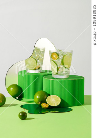 A glass containing fresh lemon slices and ice on a green platform. The image is reflected in the mirror. Pastel color background. Fresh space with fruits rich in vitamin C. 109918985
