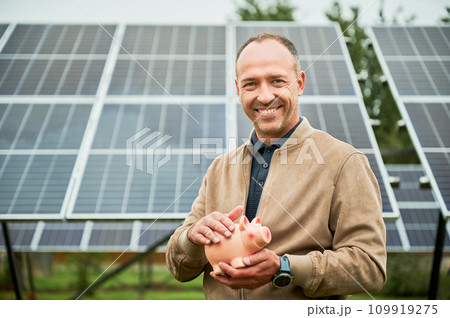 Man with piggy bank in hands smiling to camera. Young man happy about his investments in alternative energy. Enthusiastic investor glad about earnings he got. Man with piggy bank in hands smiling to camera. Young man happy about his investments in alternative energy. Enthusiastic investor glad about earnings he got. 109919275