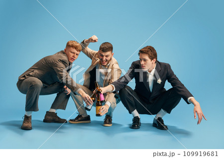 Men in stylish suits, friends attending party, drinking beer against blue background. Wedding day. Different emotions 109919681