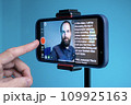 A young man shoots a video on a smartphone while reading text from a teleprompter on a screen 109925163